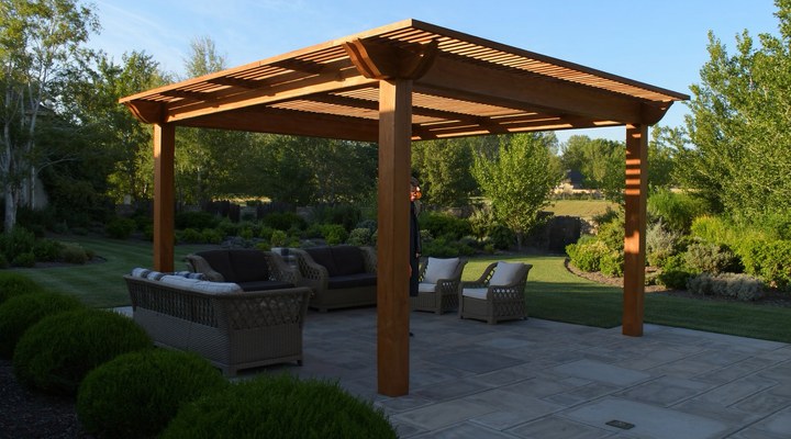 A freestanding cedar pergola with slatted roof over a stone patio in a manicured backyard, with lounge seating beneath — example of pergola construction work by Ponderosa Builders LLC.