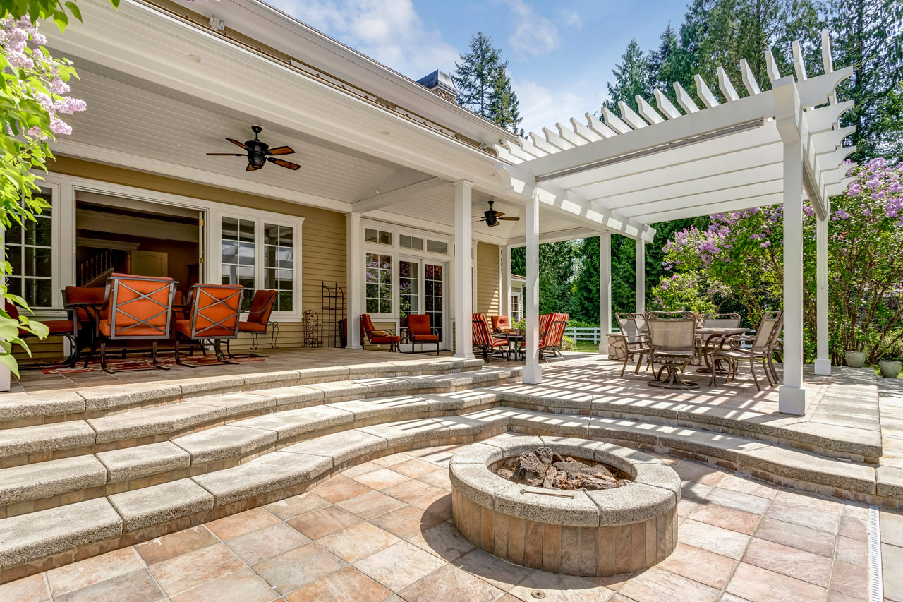 An outdoor living space under a white pergola with a stone patio and circular fire pit, perfect for pergola construction and patio design.